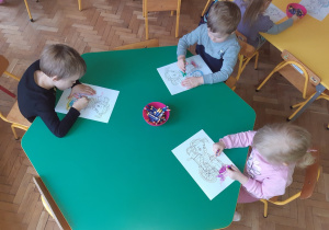 1 dziewczynka i 2 chłopców siedzi przy zielonym sześciokątnym stole i koloruje obrazki przedstawiające dziewczynkę na wózku inwalidzkim.