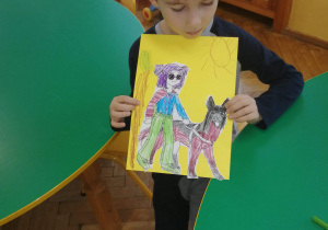 Na zdjęciu chłopiec z grupy fioletowej siedzi przy zielonym okrągłym stoliku i prezentuje prace na żółtym kartonie niedowidząca dziewczynka z psem przewodnikiem
