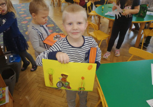 Na zdjęciu chłopiec z grupy fioletowej stoi przy zielonym kwadratowym stoliku i prezentuje prace na żółtym kartonie chłopiec na wózku inwalidzkim