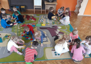 Dzieci siedzą na dywanie w grupach czteroosobowych i układają rzeczy z figur geometrycznych