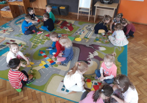 Dzieci siedzą na dywanie w grupach czteroosobowych i układają rzeczy z figur geometrycznych