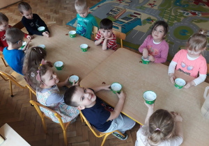 Na zdjęciu dzieci siedzą przy połączony stołach z kubeczkami po serkach wiejskich, na stole leży rzeżucha do siania