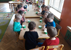 Na zdjęciu dzieci siedzą przy połączony stołach z kubeczkami po serkach wiejskich, na stole leży rzeżucha do siania