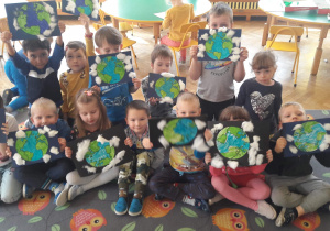 Dzieci siedzą na szarym dywanie w sowy trzymają w rękach pracę plastyczną z okazji Dnia Ziemi-Planeta Ziemia pomalowana kredkami na czarnym kartonie z tyłu pozostałe dzieci siedzą przy stolikach