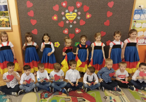 Dzieci z grupy pomarańczowej stoją na tle dekoracji z serc z przodu siedzą chłopcy ubrani w koszule z tyłu dziewczynki ubrane w suknie hiszpańskie