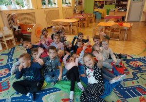 Na zdjęciu dzieci z grupy czerwonej siedzą na dywanie i pozują z okazji Dnia Przedszkolaka za plecami dzieci stoły oraz szafki