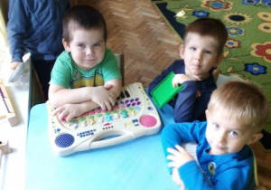 Trzech chłopców siedzi przy stoliku, na którym leży zabawkowe pianino. Za nimi stoi czwarty chłopiec. W tle widać nogi innych dzieci.