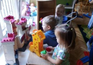 Dwie dziewczynki i jeden chłopiec bawią się w kąciku lalek. Jedna dziewczynka wkłada klocki do plastikowej żyrafy, chłopiec bawi się zamkiem. Druga dziewczynka bierze z szafki zabawkowe naczynia.