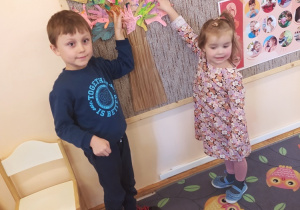 Na zdjęciu dziewczynka i chłopiec pokazują na tablicy wykonany przez siebie szablon własnej dłoni, który stanowi koronę papierowego drzewa.