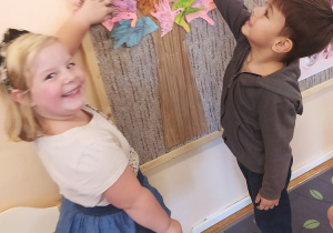 Dziewczynka i chłopiec wskazują na tablicy własnoręcznie zrobiony szablon swojej dłoni, który tworzy koronę papierowego, grupowego drzewa.