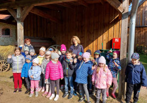 Na zdjęciu dzieci z grupy czerwonej z wychowawcami na tle drewnianej stodoły w skansenie