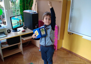 Na zdjęciu chłopiec stoi na dywanie i bawi się niebieską kostką, chłopiec podnosi jedą rękę do góry