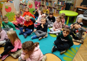 Na zdjęciu dzieci z grupy zielonej podczas zajęć w Bibliotece Rejonowej nr 13 dzieci siedzą w czytelni na kolorowym dywanie wokół półki z książkami
