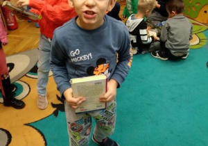 Na zdjęciu chłopiec z grupy zielonej trzyma w rękach książkę wokół półki z książkami