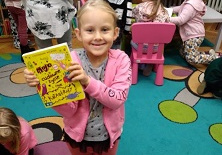 Na zdjęciu dziewczynka z grupy zielonej trzyma w ręku żółtą książkę, dziewczynka jest uśmiechnięta