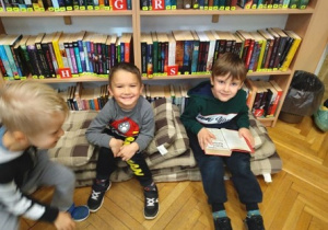 Na zdjęciu trzej chłopcy z grupy zielone siedzą na poduszka z książkami z tyłu za nimi wysoki regał z książkami