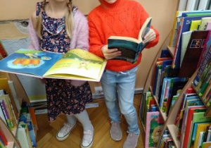 Na zdjęciu dwie dziewczynki z grupy zielonej podczas oglądania książeczek, dziewczynki stoją pomiędzy dwoma półkami z książkami