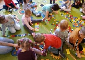 Dzieci siedzą na dywanie i układają z plastikowych figur geometrycznych pociąg.