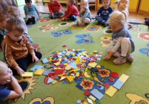 Dzieci siedzą na dywanie w kole. Chłopiec i dziewczynka siedzą w środku koła i wybierają figury geometryczne.