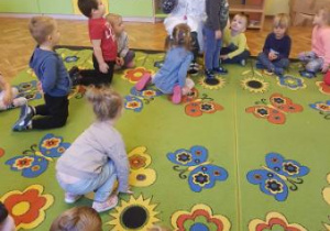 Dzieci siedzą na dywanie. Nauczycielka przebrana w kitel lekarski siedzi na krzesełku, przed nią stoi chłopiec. Nauczycielka za pomocą zabawkowego stetoskopu pokazuje dzieciom badanie lekarskie.