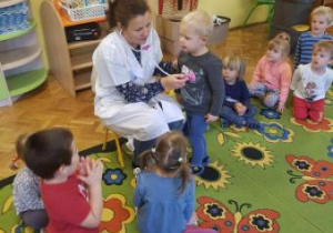 Nauczycielka przebrana w fartuch lekarski siedzi na krzesełku. Przy pomocy zabawkowego stetoskopu pokazuje dzieciom na stojącym przed nią chłopcu sposób osłuchiwania serca przez lekarza.