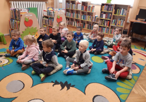 Dzieci siedzą na dywanie i słuchają wierszy czytanych przez Panią bibliotekarkę. Za dziećmi widać regały z książkami.