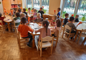 Dzieci siedzą przy stolikach. Rozczesują szczotkami pomarańczowo-zielone sznurki, z których będą robić marchewki.