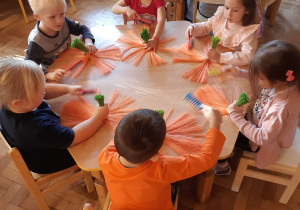 Pięć dziewczynek i pięciu chłopców siedzi przy stolikach, formują z pomarańczowych sznurków wachlarzyk.
