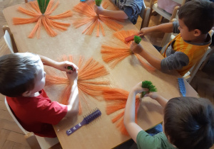 Czterech chłopców i jedna dziewczynka siedzą przy stole. Rozczesują szczotkami pomarańczowo-zielone sznurki.