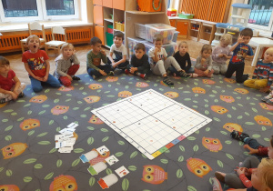Dzieci siedzą w kole. Na środku dywanu rozłożona jest plansza podzielona na kwadraty, a na niej rozłożone rysunki owoców. Obok planszy położone są trzy słoiki oznaczone cyframi: 1, 2 3 oraz kartoniki z narysowanymi strzałkami.
