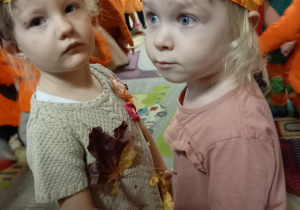 Dwie dziewczynki w jesiennych opaskach na głowie trzymają się za ręce - tańczą w rytm jesiennej piosenki.