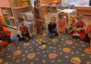 Dzieci w przebraniach jesiennych siedzą na dywanie, jedna dziewczynka stoi w środku koła. W tle widać kącik lalek oraz pudełka z klockami.