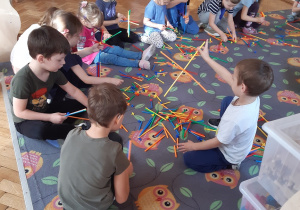 Dzieci siedzą na dywanie w sowy i budują z kolorowych klocków - słomek wymyślone budowle.