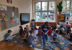 Dzieci siedzą na dywanie i budują z plastikowych, kolorowych słomek - klocków przestrzenne budowle. Jeden chłopiec idzie po dywanie.