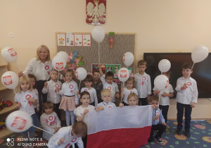Dzieci i nauczycielka w strojach galowych i kotylionach ustawieni są na tle tablicy. Niektóre dzieci trzymają białe balony z namalowanym godłem Polski. Troje dzieci trzyma biało-czerwoną flagę Polski.