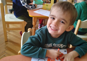 Na zdjęciu chłopiec siedzący przy stole koloruje rysunek misia chłopiec uśmiecha się