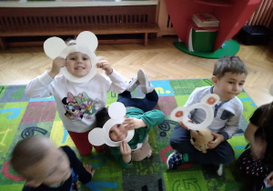 Na zdjęciu dzieci siedząe na dywanie dzieci mają białe maski w kształcie misia