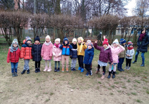 Dzieci z grupy zielonej stoją w ogrodzie ubrane w kurtki i czapki dzieci są ustawione podczas zbiórki