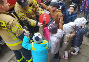 Dzieci stoją prze wozie strażackim. Strażak zakłada dziewczynce kask strażacki.