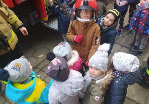 Dzieci stoją przy wozie strażackim. Podnoszą ręce z chęcią zadania pytania strażakom. Jeden chłopiec pozuje do zdjęcia w kasku strażackim.