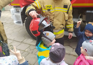 Dzieci słuchają opowieści strażaków o ich pracy. Strażak zakłada chłopcu kask.