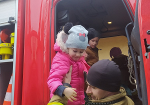 Dzieci oglądają od środka wóz strażacki. Strażak pomaga dziewczynce w zejściu z wozu.