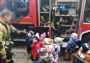 Dzieci stoją przy wozie strażackim. Słuchają strażaków opowiadających o swojej pracy.