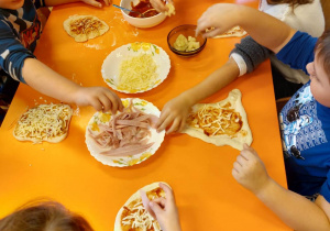 Na zdjęciu dzieci z grupy czerwonej, dzieci siedzą przy pomarańczowym stoliku dzieci sięgają po produkty na pizze