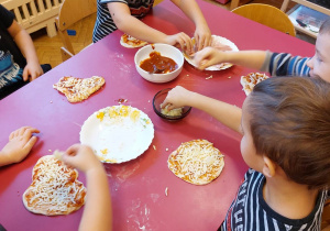 Na zdjęciu trzech chłopców przy różowym stole nakładają ser na ciasto do pizzy