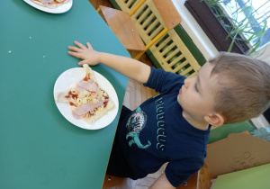 Chłopiec siedzi przy stole, na którym stoi talerzyk ze zrobioną przez chłopca pizzą.