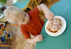 Chłopiec w czerwonej bluzce siedzi przy stole. Własnoręcznie robi swoją pizzę.
