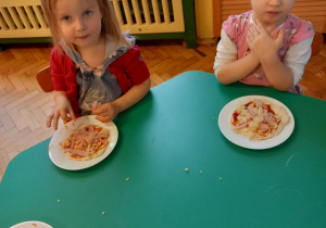 Dwie dziewczynki siedzą przy stole. Przed dziewczynkami stoją talerze z wykonanymi przez nie pizzami.