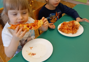 Dziewczynka i chłopiec siedzą przy stoliku. Jedzą własnoręcznie zrobioną pizzę.