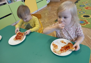Dziewczynka i chłopiec siedząc przy stoliku, jedzą własnoręcznie zrobioną pizzę.